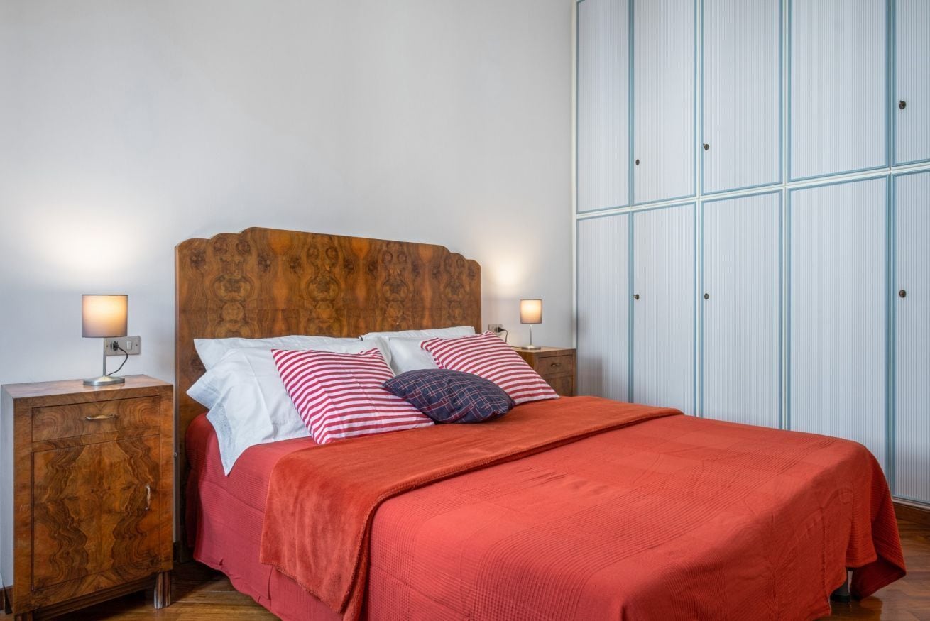 Una cama bien hecha con un cabecero de madera, dos mesitas de noche con lámparas y un amplio armario con tiradores verticales.