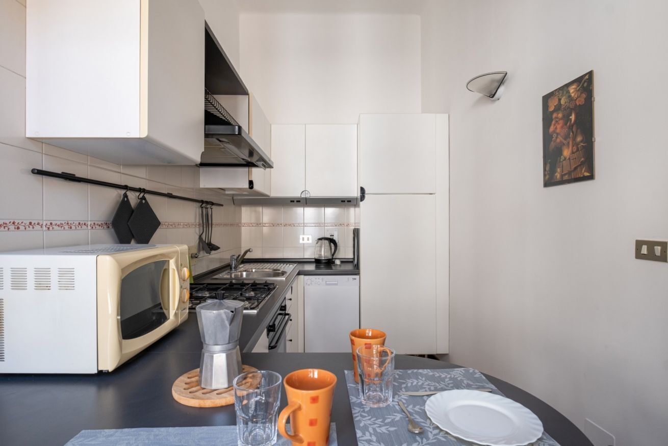 Una cocina compacta con gabinetes blancos, un microondas, una hornilla y un lavamanos. Hay dos tazas naranjas y un vaso en la encimera.