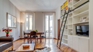 A modern living room with a wooden floor, a dining area with a table and chairs, a sofa, a coffee table, a TV on a white cabinet, and a ladder.