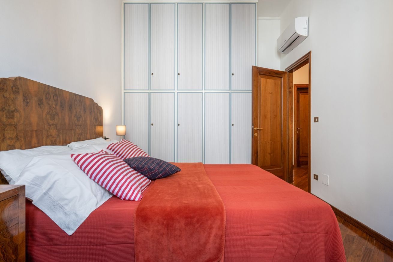 A bedroom with a wooden headboard, a bed with red bedding, striped pillows, and a red throw blanket. There is a wardrobe with light blue panels and a wooden door.