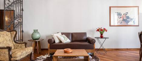 Un salon avec un canapé en cuir marron, un fauteuil à motifs, une table basse en bois, un vase vert et un tableau encadré accroché au mur.
