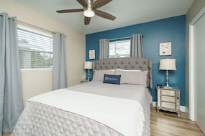 A bedroom with a large bed, a ceiling fan, a nightstand with a lamp, and a window with curtains.