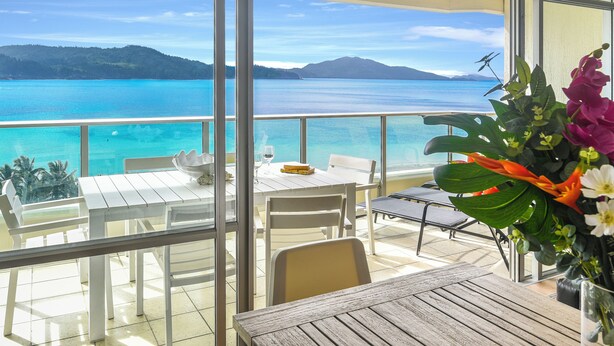 A balcony with a table and chairs, overlooking a turquoise sea and distant mountains.