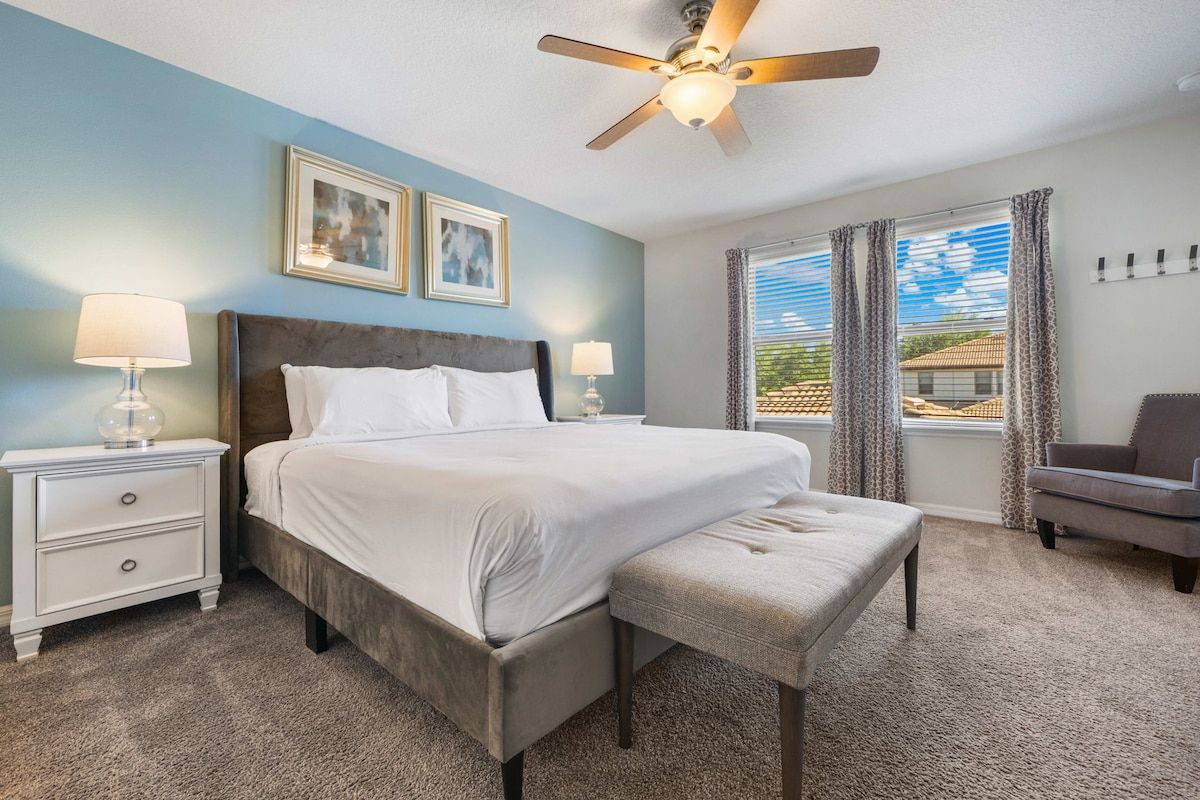 A bedroom with a bed, two bedside tables, a chair, a ceiling fan, and a window with curtains.