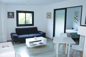A modern, minimalistic room with a black sofa, a white coffee table, a small dining table with chairs, and a bed with a blue headboard. - Charming little house in a green setting (Ravine des Cabris)