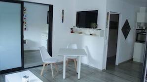 Habitación moderna y minimalista con una mesa blanca y dos sillas, un televisor de pantalla plana sobre la mesa, y una cocina visible a través de una puerta abierta.