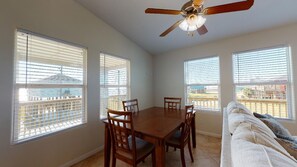 A dining area with a wooden table and chairs, a ceiling fan, and large windows with blinds.