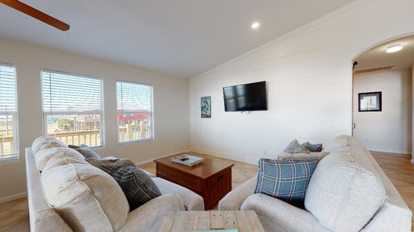 A living room with a sofa set, a coffee table, a flat-screen TV mounted on the wall, and large windows with blinds.