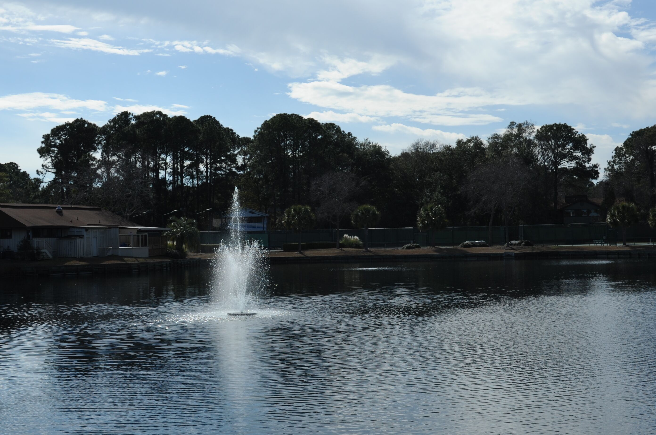 A tranquil lake with a fountain in the center, surrounded by trees and a house in the background.