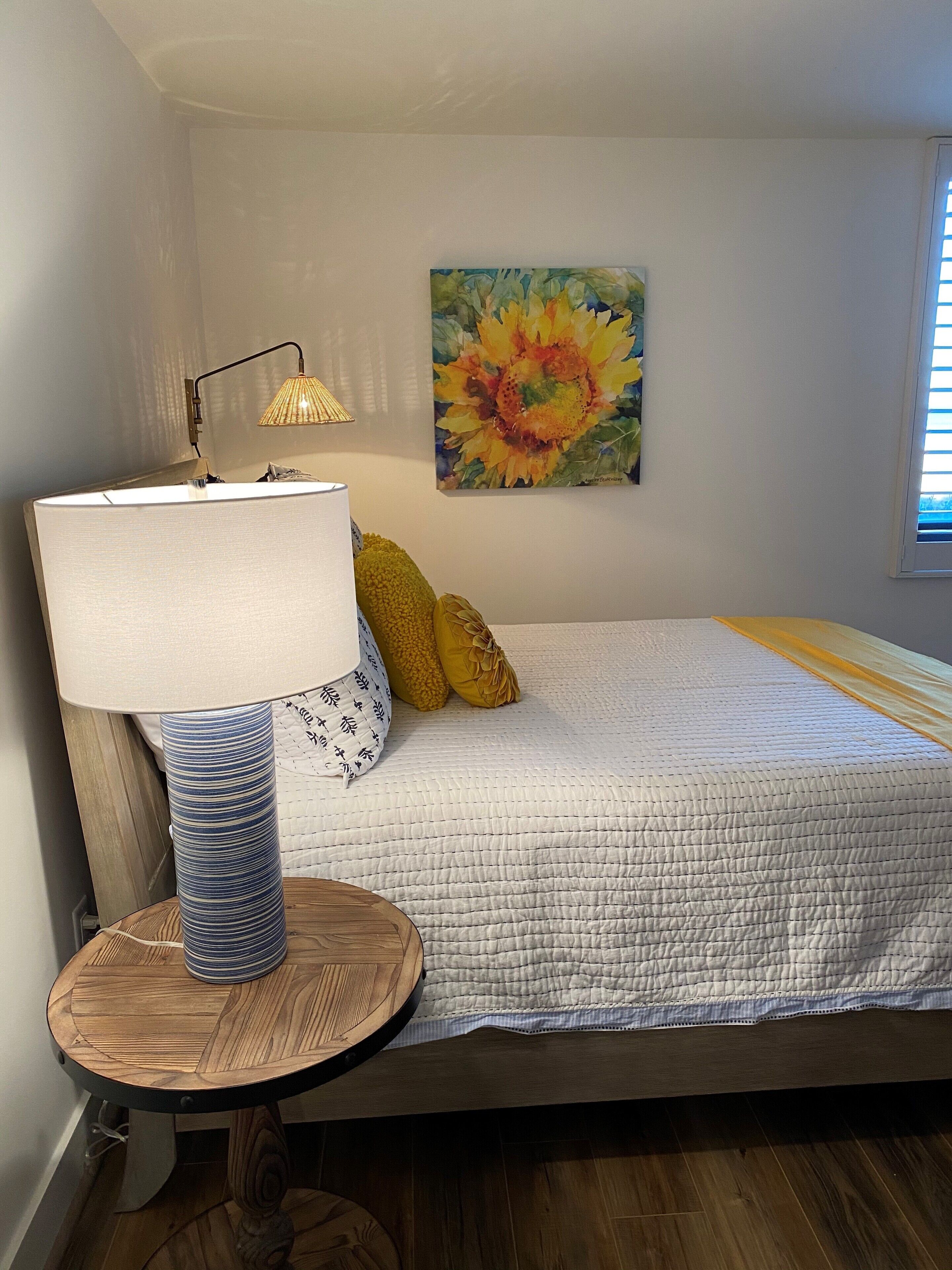 A bedroom with a bed, a round wooden table with a lamp, a painting on the wall, and a window with blinds.
