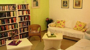 A cozy living room with a bookshelf, a wicker chair, a white sofa with floral cushions, and a small table with candles. - Gioma Hotel (Agios Vasileios)