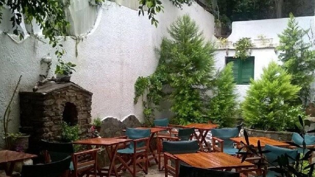 Une terrasse extérieure avec des tables et des chaises en bois, entourée de verdure et d’une structure en pierre.