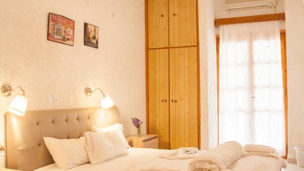 A bedroom with a bed, wooden wardrobe, bedside lamps, and a window with curtains. - Gioma Hotel (Agios Vasileios)