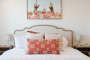 Una cama bien hecha con sábanas blancas, un cojín decorativo estampado y un cuadro enmarcado de un paisaje desértico encima.