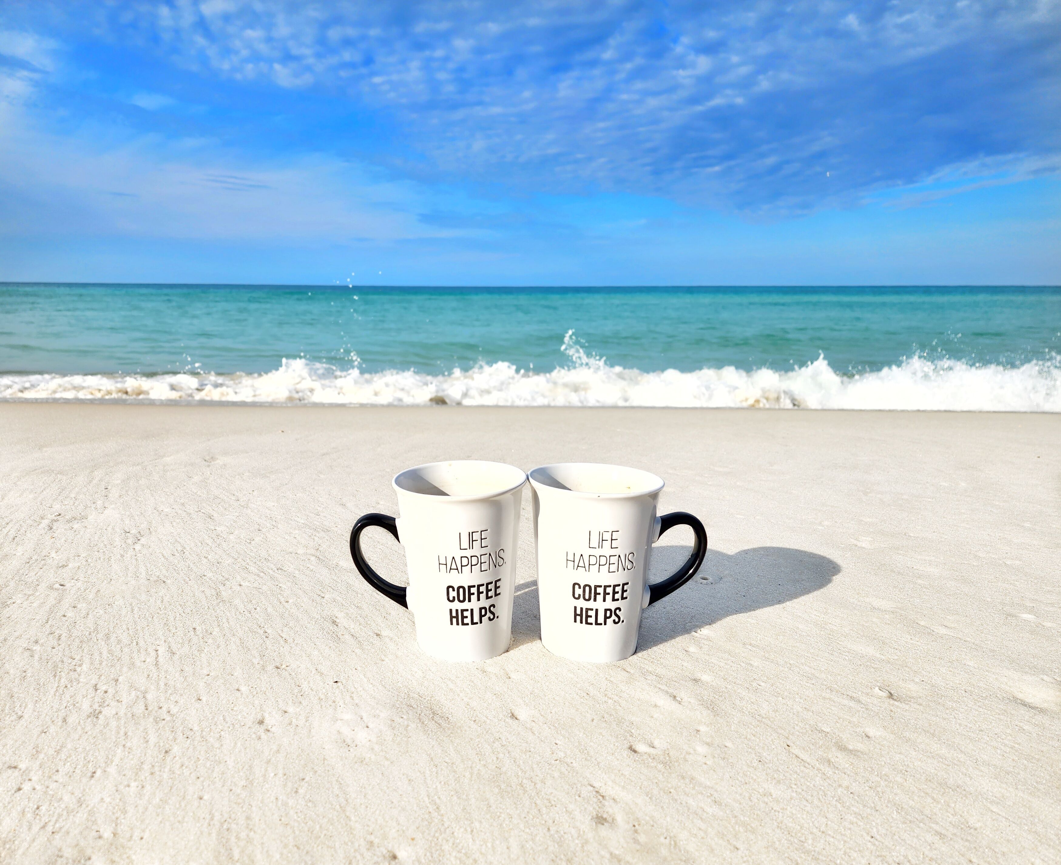 Two white coffee mugs with black handles and text on a sandy beach with ocean waves in the background.