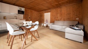 Une cuisine moderne avec des meubles blancs, un four à micro-ondes et une table à manger en verre, entourée de chaises blanches. Le salon comprend un canapé blanc et une petite table sur laquelle est posé un jeu de société.