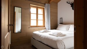Une chambre avec un plancher en bois, un lit recouvert de draps blancs, une fenêtre avec des rideaux et un miroir au mur.