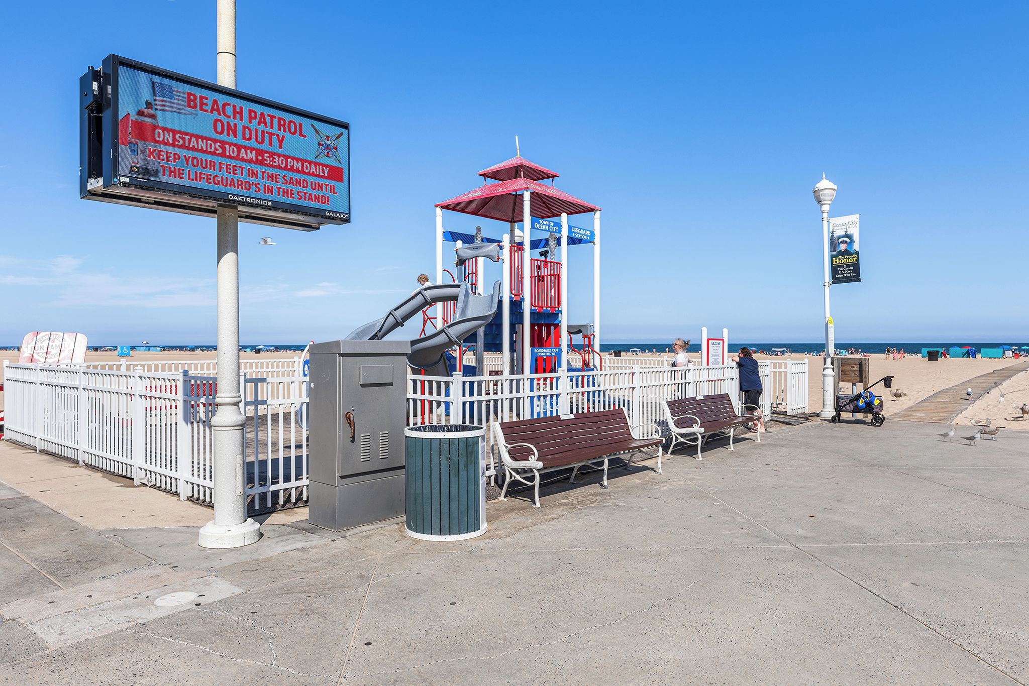 A beachside lifeguard station with a red and blue structure, a bench, a trash can, and a signboard.