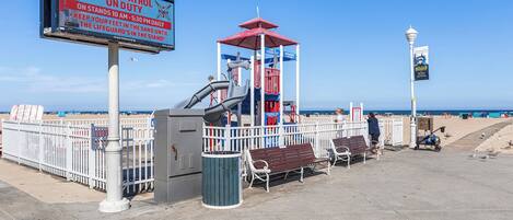 A beachside lifeguard station with a red and blue structure, a bench, a trash can, and a signboard.