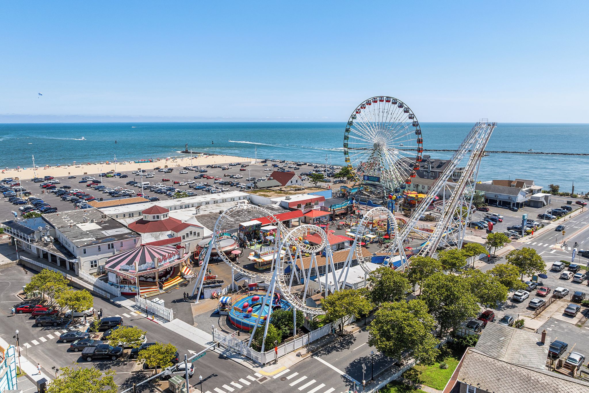 An aerial view of a coastal amusement park with a large Ferris wheel, roller coasters, and a parking lot.