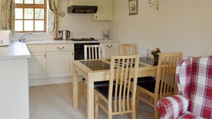 A compact kitchen with white cabinets, a dining table with chairs, a red plaid sofa, and a window with curtains. - 2 bedroom accommodation in Sidbury, near Sidmouth (Sidbury, near Sidmouth)