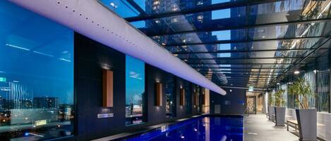Un área de piscina interior moderna con techo de cristal, vistas a la ciudad y un diseño elegante.