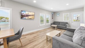 A modern living room with a grey sofa, a wooden coffee table, and a dining table with chairs. There are large windows with blinds, a painting on the wall, and a door with a view of the outside. - Spacious 3BR apartment 213B near Boardwalk and the Beach (Ocean City)
