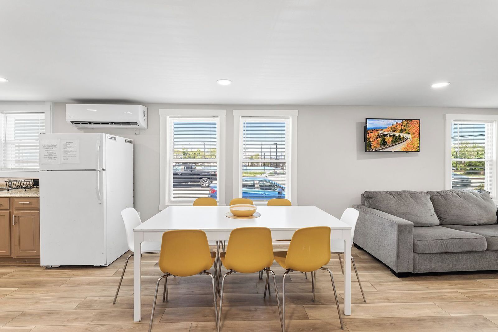 A modern kitchen with a white refrigerator, a dining table with four yellow chairs, and a gray sofa.