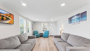 Een moderne woonkamer met twee grijze banken, een blauwgroene fauteuil en een kleine tafel. Er zijn grote ramen met zonwering, een deur en een aan de muur bevestigde televisie waarop een schilderachtige afbeelding staat.