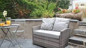 A wicker sofa with cushions, a metal table with a glass of orange juice, and a metal chair. - 2 bedroom accommodation in Bryncrug, near Tywyn (Bryncrug, near Tywyn)