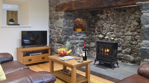 A living room with a stone wall, a television, a wooden coffee table with fruit and wine, and a fireplace. - 2 bedroom accommodation in Bryncrug, near Tywyn (Bryncrug, near Tywyn)