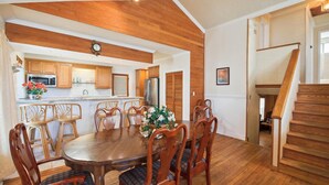 A dining area with a wooden table and chairs, a kitchen in the background, and a wooden staircase.