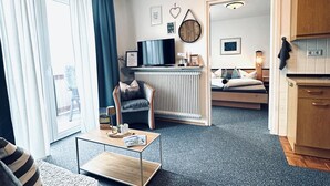 Un soggiorno moderno con un divano, un tavolino e una TV. È presente una zona cucina con pensili e un tavolo da pranzo. La stanza ha un'ampia finestra con tende.