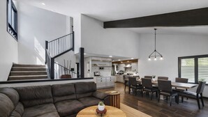 Ein modernes Wohnzimmer mit einem dunkelbraunen Ecksofa, einem runden Couchtisch und einem Essbereich mit Holztisch und -stühlen. Im Hintergrund ist eine Küche mit weißen Schränken und einer schwarzen Treppe zu sehen.