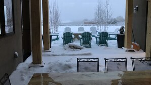 A covered outdoor seating area with snow-covered chairs and tables, wooden beams, and a view of a snowy landscape.