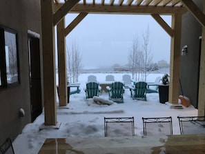 Uma área de estar ao ar livre coberta, com cadeiras e mesas cobertas de neve, vigas de madeira e vista para uma paisagem nevada.