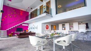 Un intérieur moderne avec un mur rose vif, un escalier, un coin repas doté d’une table en verre et une cuisine aux meubles blancs.