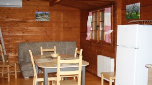 Interior de una cabaña de madera con una mesa de comedor, sillas, un sofá, un refrigerador y una ventana con cortinas.