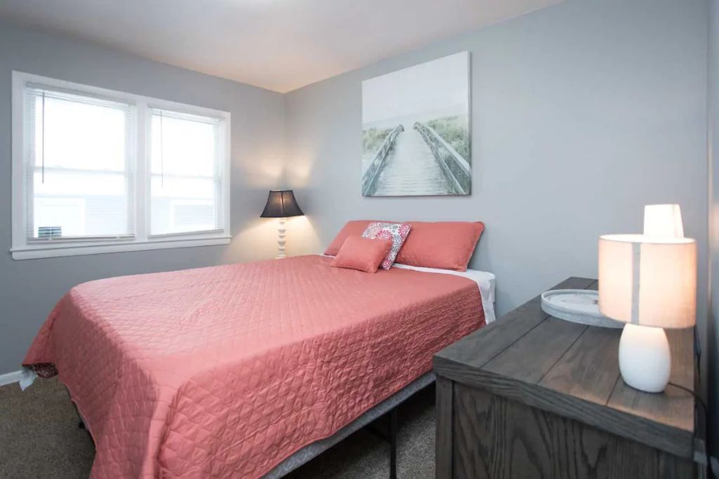 A bedroom with a bed, a nightstand, a lamp, a window, and a wall art piece.