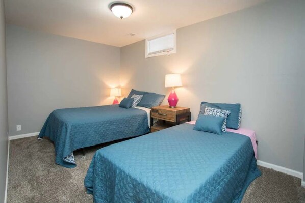 A bedroom with two beds, each with a blue bedspread and matching pillows, a wooden nightstand with a lamp, and a window above the beds. - ❤️ Unbeatable group travel retreat ❤️ (Omaha)