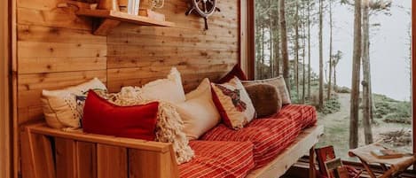 A cozy cabin interior with a wooden bench and sofa, a table with books, and a view of the forest through a large window.
