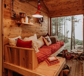 A cozy cabin interior with a wooden bench and sofa, a table with books, and a view of the forest through a large window. - Perch small · Treehouse-Eagles Perch over the water (Port Angeles)