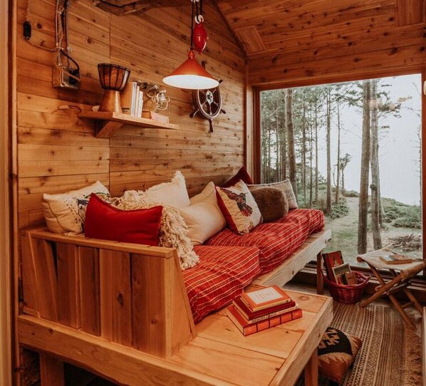 A cozy cabin interior with a wooden bench and sofa, a table with books, and a view of the forest through a large window.