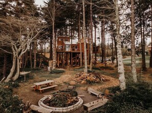 A cabin in a forest with a fire pit, picnic tables, and a wooden deck.
