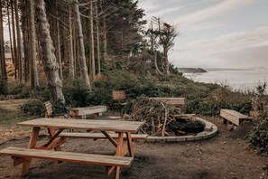 A rustic outdoor seating area with picnic tables and a fire pit, surrounded by tall trees and overlooking a body of water.