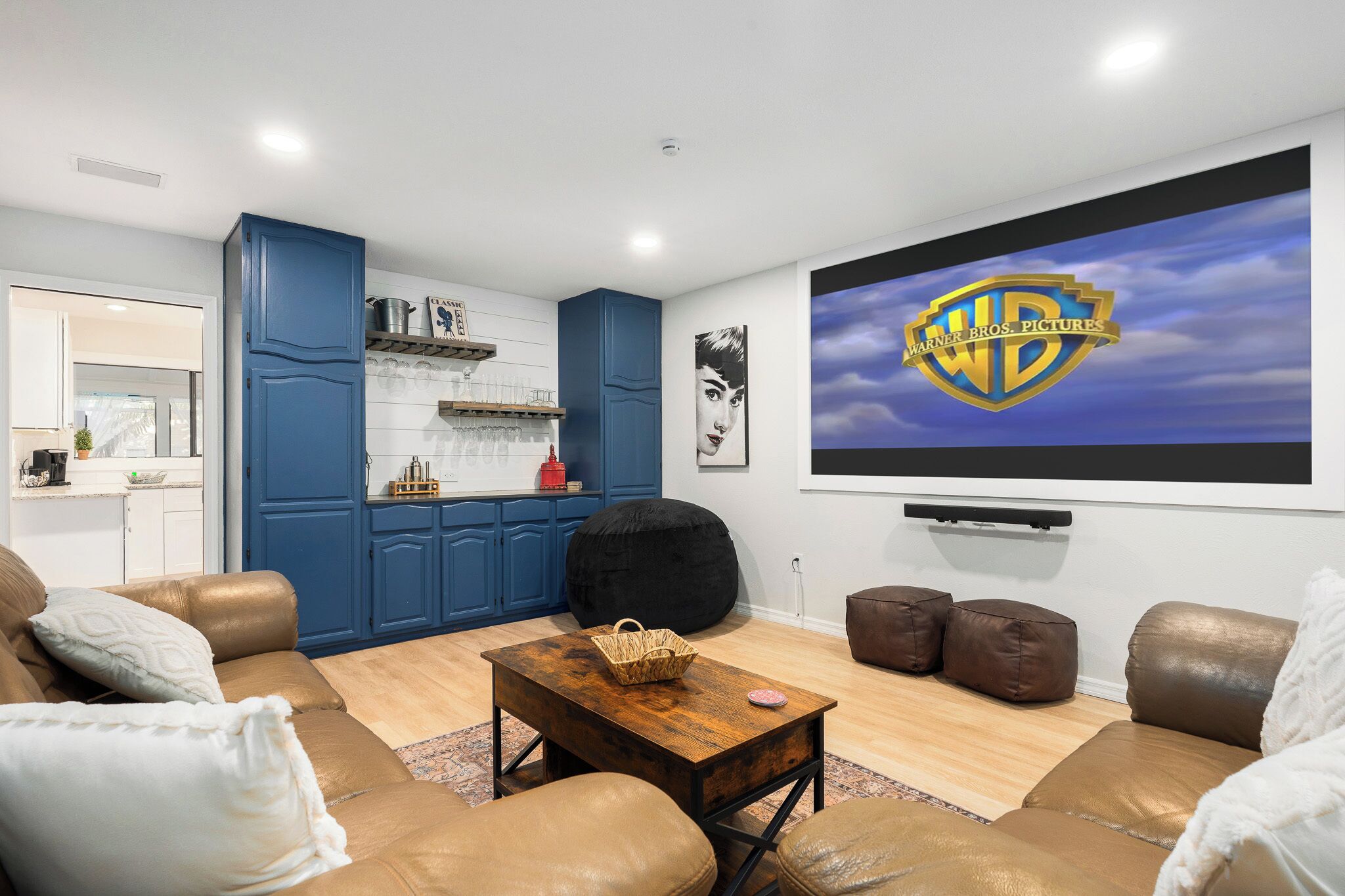 Olohuoneessa on suuri näyttö, jossa näkyy Warner Bros. Pictures -elokuvayhtiön logo. Huoneessa on myös ruskea nahkasohva, puinen sohvapöytä ja keittiöalue, jossa on siniset kaapit.