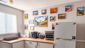 Et køkken med køleskab, ovn, kaffemaskine og et vindue med persienner.