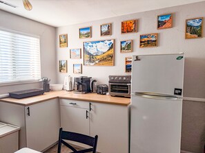 A kitchen with a refrigerator, oven, coffee maker, and a window with blinds.