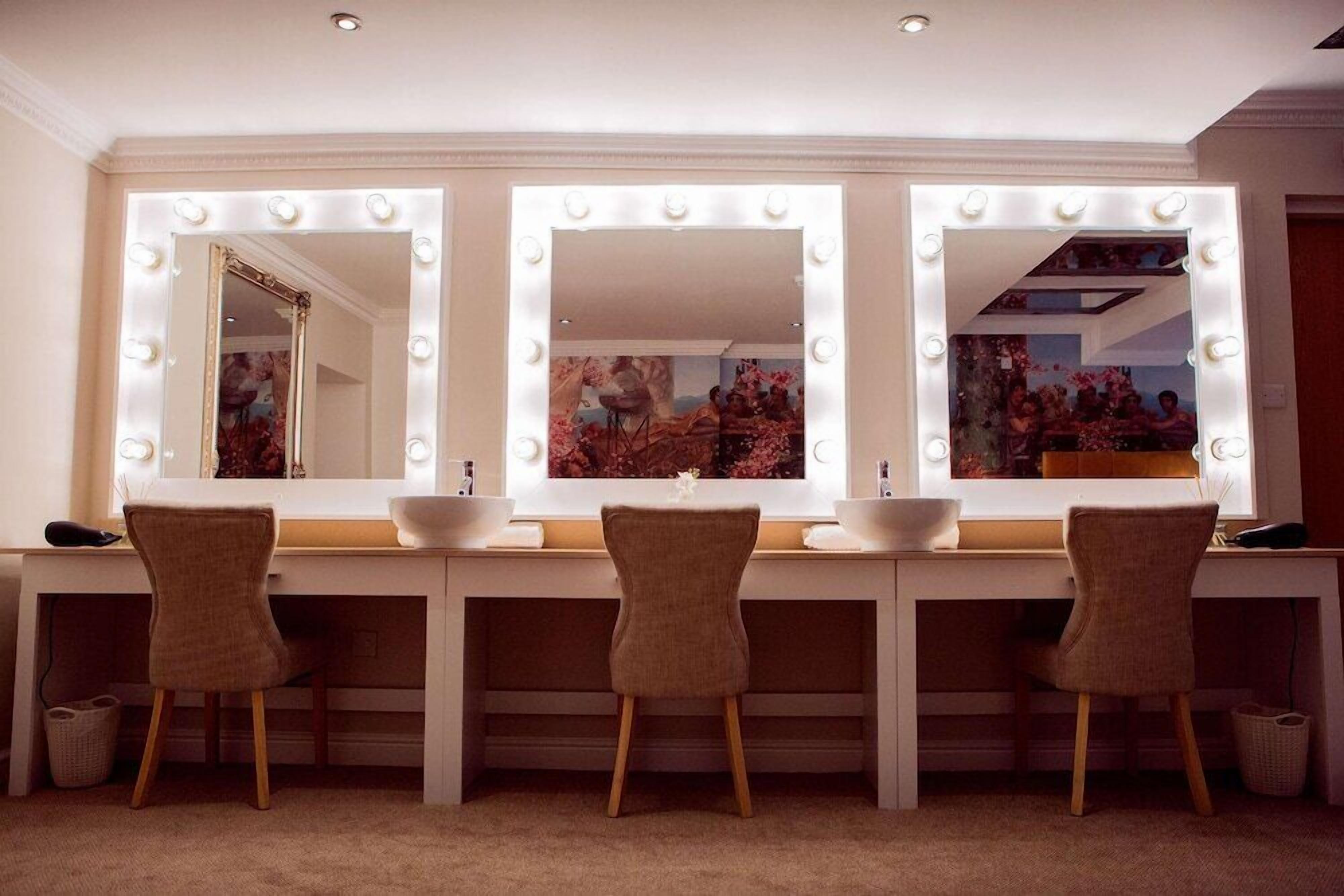 A vanity area with three mirrors, each surrounded by lights, and a white countertop with a sink and chair.