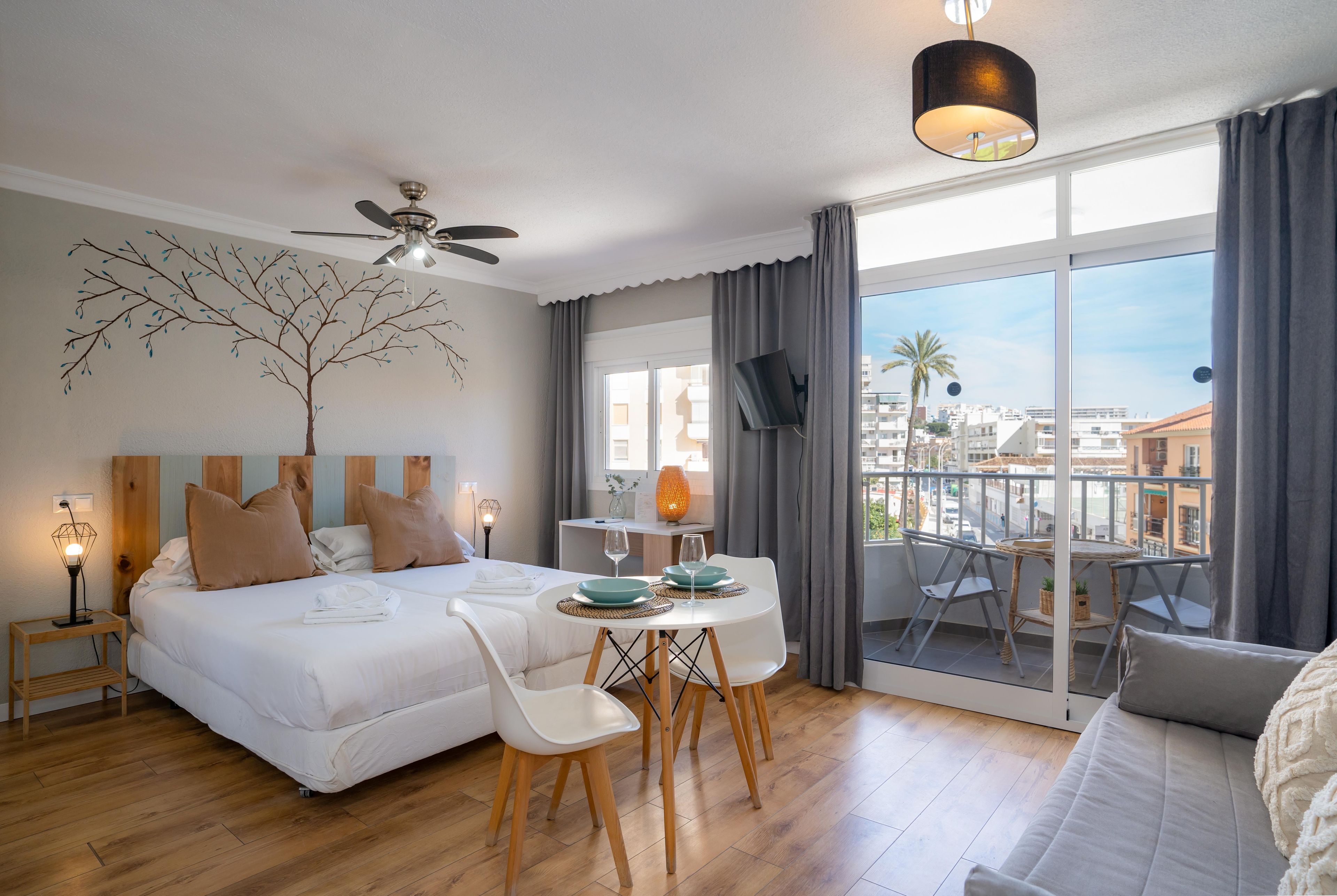 A hotel room with a bed, a dining table with chairs, a TV, and a balcony with a view of the city.
